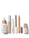 A collection of ILIA beauty products, including foundation, concealer, mascara, lip pencil, and blush, arranged neatly on a white background.