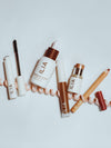 ILIA beauty products scattered on a white surface, including, Mascara, Skin Tint, Concealer, Multi-Stick, and Lip Liner.